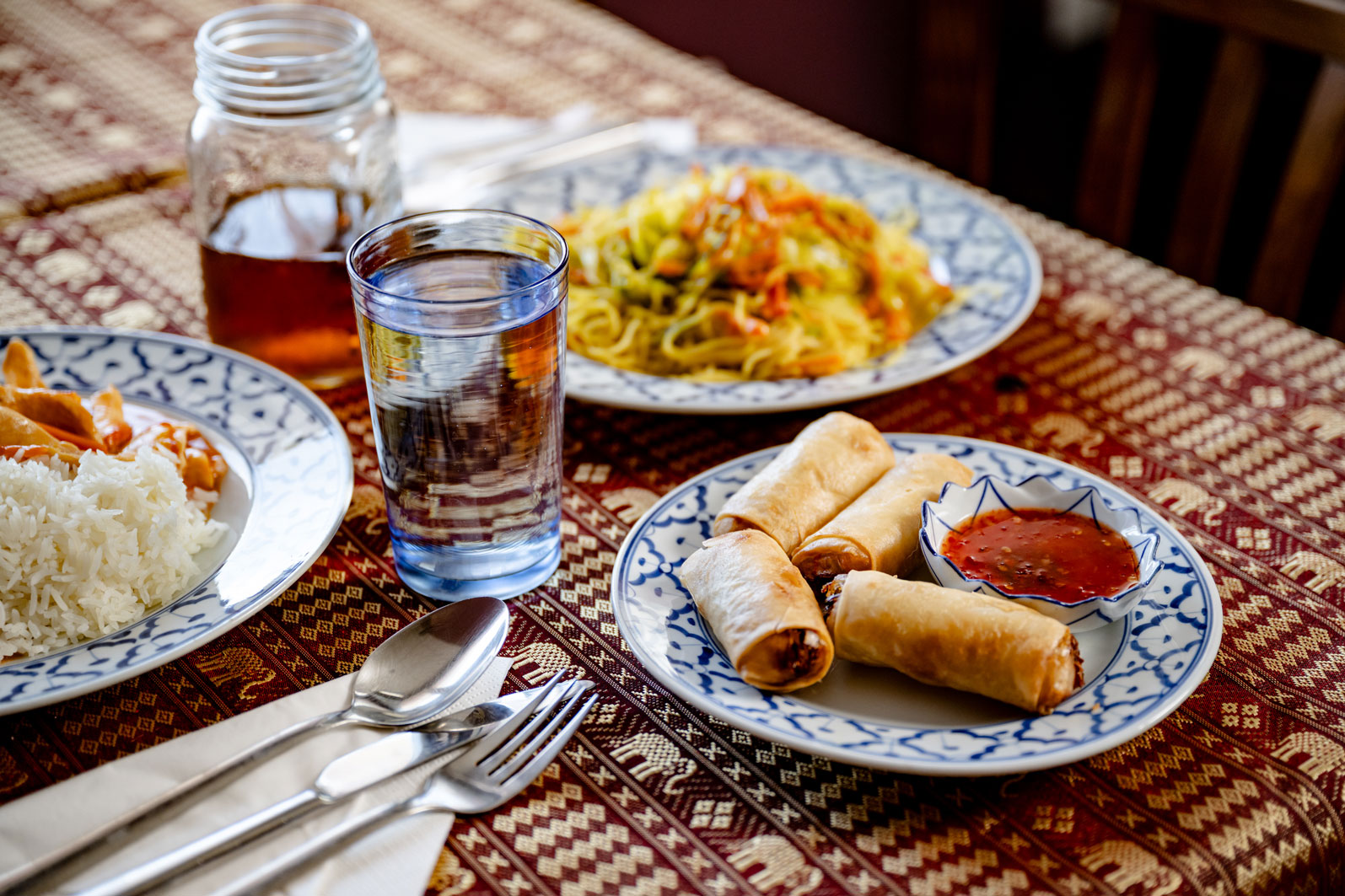 Thailändische Gerichte mit Frühlingsrollen und Getränken Verschiedene thailändische Gerichte auf traditioneller Tischdecke, darunter Frühlingsrollen mit süsser Chilisauce, Reis mit Curry, gebratene Nudeln und Getränke im Einmachglas und Glas.