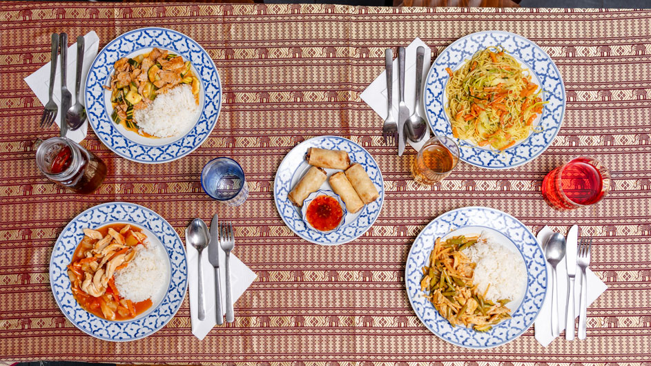 Traditionelles thailändisches Mittagessen mit Reisgerichten, Frühlingsrollen und Getränken Ansicht von oben auf ein traditionelles thailändisches Essen auf einer gemusterten Tischdecke mit vier Tellern Reis und verschiedenen gebratenen Gerichten, einem Teller Frühlingsrollen mit Dip-Sauce und Gläsern mit Getränken.