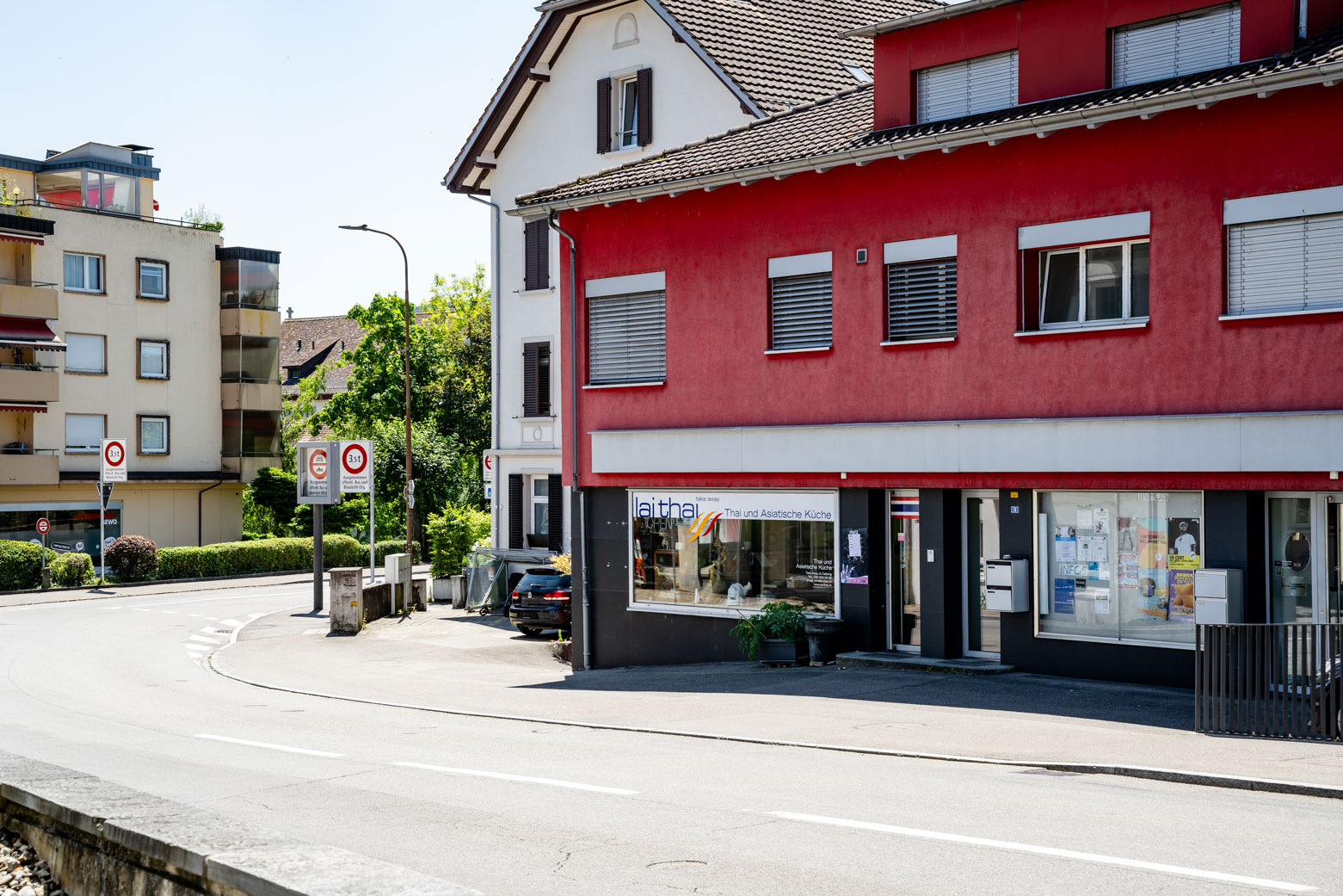 Strassenansicht des Lai Thai Take Away Restaurants im Erdgeschoss eines roten Eckgebäudes mit sichtbarer Beschilderung und angrenzenden Geschäften.